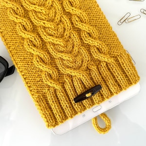 May include: A mustard yellow knitted tablet case with a cable knit pattern and a brown button closure. The case is lying on a white surface with a tablet inside.