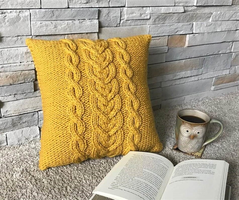 Pu&ograve; includere: Un cuscino in maglia giallo senape con un motivo a treccia. Il cuscino &egrave; appoggiato su una superficie di pietra grigia con un libro e una tazza di caff&egrave;.
