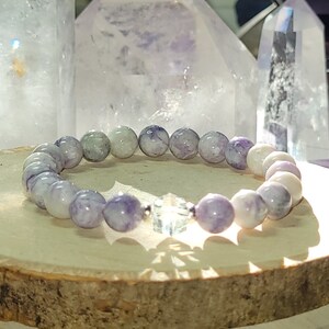 May include: A purple and white gemstone bracelet with a clear crystal charm. The bracelet is made of round beads and is sitting on a wooden surface.