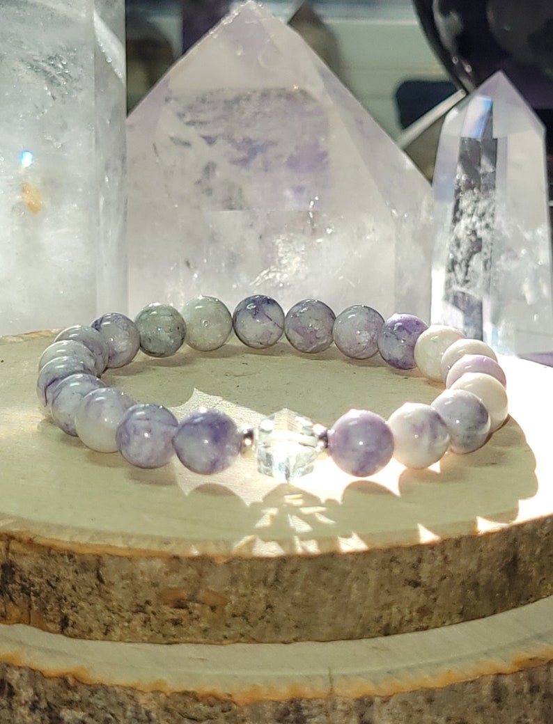 May include: A purple and white gemstone bracelet with a clear crystal charm. The bracelet is made of round beads and is sitting on a wooden surface.