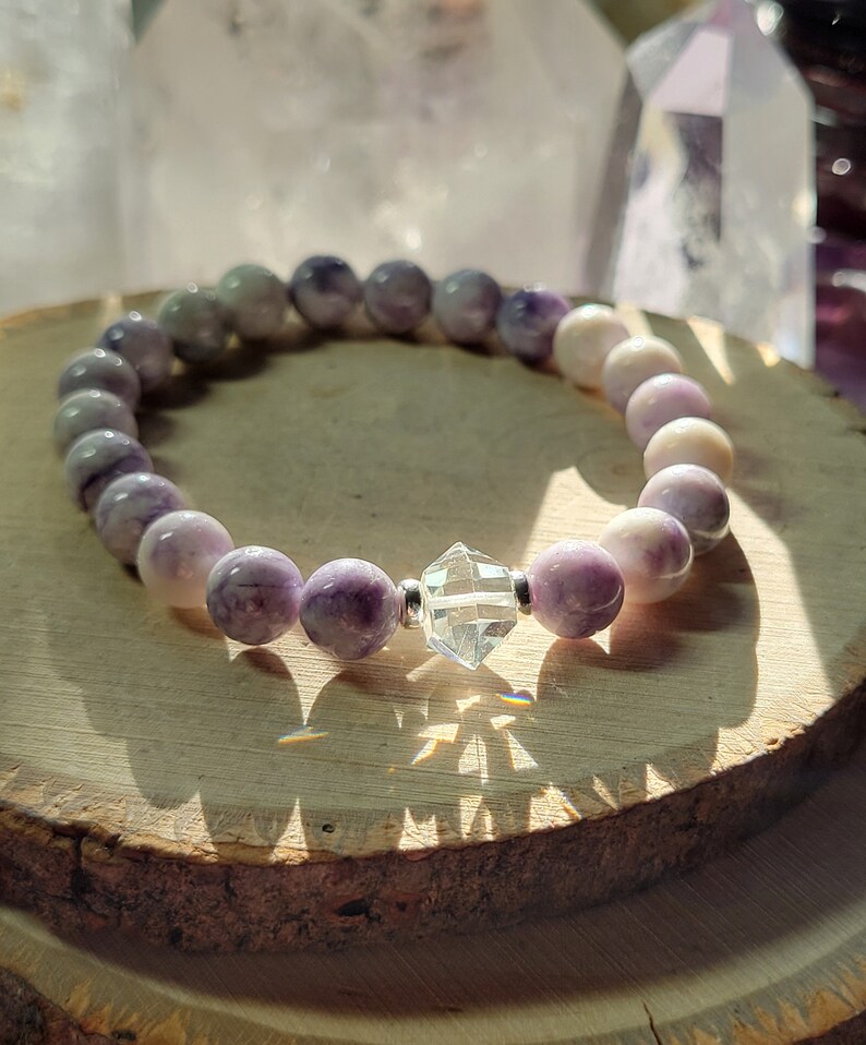 May include: A purple and white gemstone bracelet with a clear crystal charm. The bracelet is made of round beads and is strung on elastic.