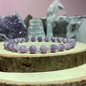 May include: A purple and clear crystal beaded bracelet on a wooden surface. The bracelet is made with faceted amethyst and clear quartz beads.