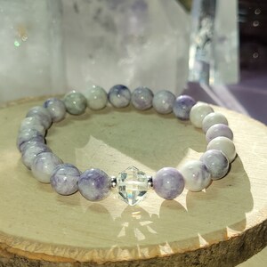 May include: A purple and white gemstone bracelet with a clear crystal charm. The bracelet is made of round beads and is strung on a silver chain.