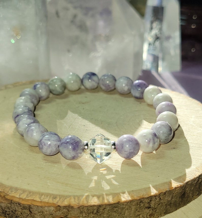 May include: A purple and white gemstone bracelet with a clear crystal charm. The bracelet is made of round beads and is strung on a silver chain.