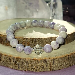 May include: A purple and white gemstone bracelet with a clear crystal charm. The bracelet is made of round beads and is strung on elastic.