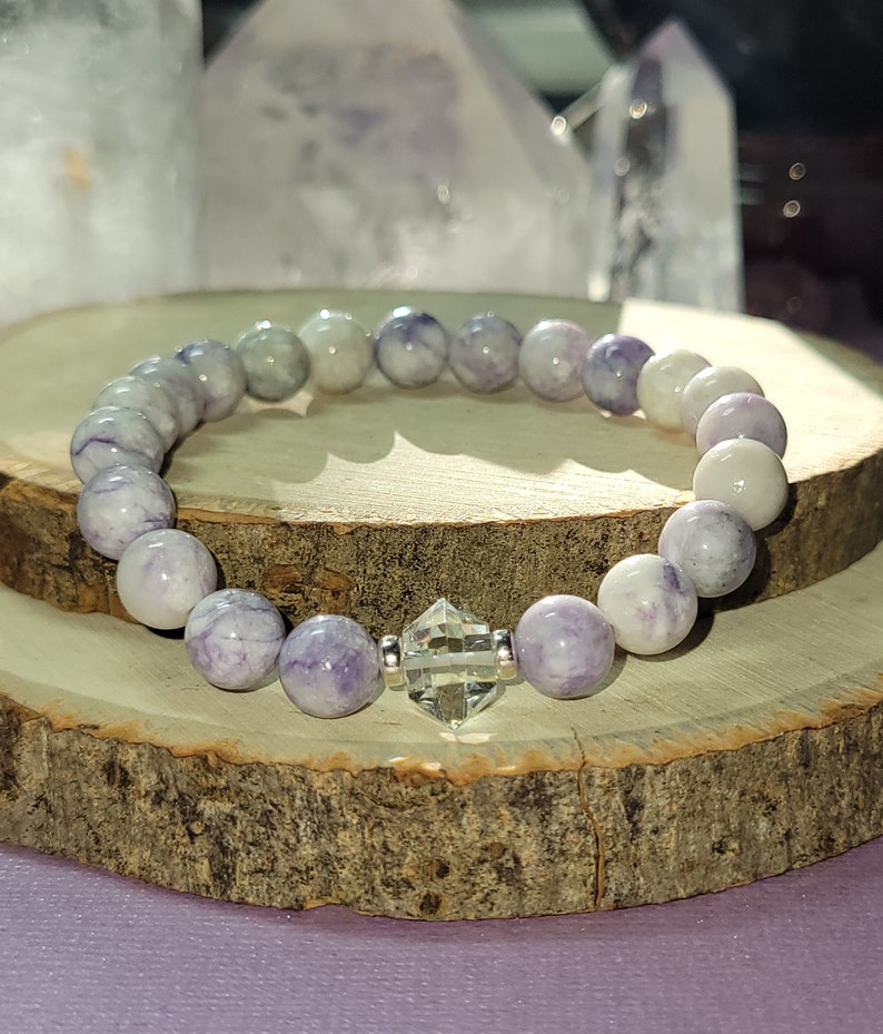May include: A purple and white gemstone bracelet with a clear crystal charm. The bracelet is made of round beads and is strung on elastic.