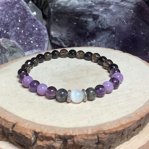 Könnte beinhalten: Ein mehrfarbiges Perlenarmband mit braunen, violetten und grauen Perlen. Das Armband hat einen silbernen Anhänger mit einem weißen Stein in der Mitte.