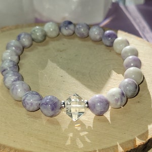 May include: A purple and white gemstone bracelet with a clear crystal charm. The bracelet is made of round beads and is strung on a silver chain.