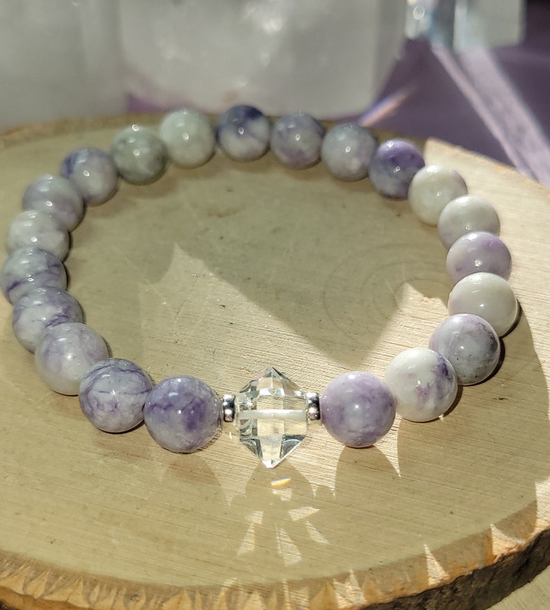 May include: A purple and white gemstone bracelet with a clear crystal charm. The bracelet is made of round beads and is strung on a silver chain.