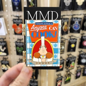 May include: A colorful enamel pin featuring a chef with the text "Anyone Can Cook!" on a blue and white tile background. The pin has gold-colored hinges and is attached to a black backing with "MMD" printed on it.