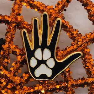 Op de afbeelding: Zwarte en gouden emaille pin in de vorm van een hand met een witte pootafdruk in het midden. De pin heeft een gouden rand en is geplaatst tegen een achtergrond van oranje glitter.