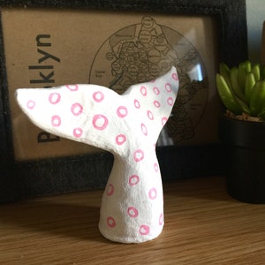 May include: White ceramic whale tail with pink polka dots. The tail is curved and has a rounded tip.