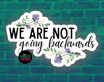 May include: A white sticker with the text "WE ARE NOT going backwards" in a mix of bold and cursive fonts. The sticker is decorated with floral elements and the logo for "the weird EMPORIUM". The background is a green brick wall.