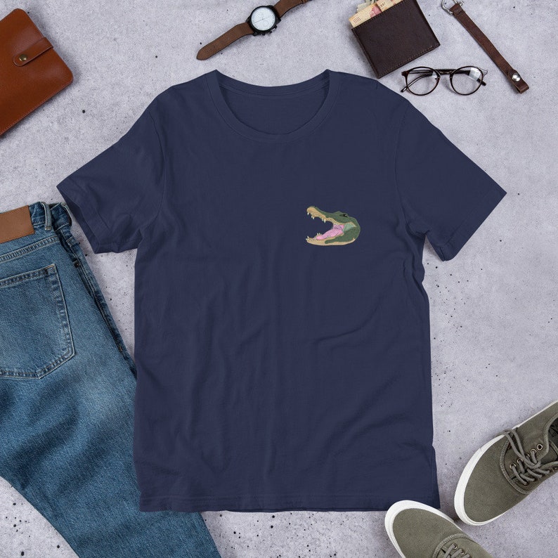 Pu&ograve; includere: Una t-shirt blu navy con una grafica di testa di alligatore verde sul petto sinistro.