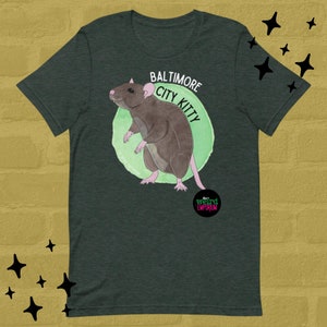 May include: A dark green t-shirt with a cartoon rat illustration and the text "Baltimore City Kitty" in a green circle. The text "The Weird Emporium" is in a black circle with pink text.