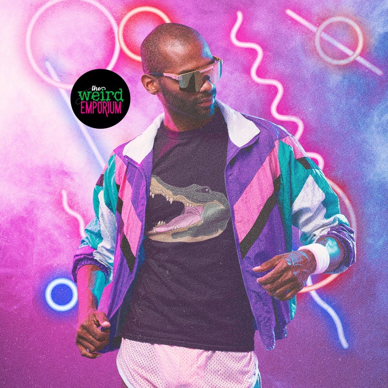 May include: A person wearing a black t-shirt with a graphic of an alligator's head. The t-shirt is layered under a purple, pink, and teal color-blocked jacket. The person is wearing sunglasses and standing in front of a pink and blue background with neon lights.