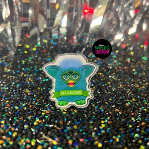 May include: A blue and green fuzzy toy with big eyes and a yellow beak. The toy is holding a green banner that says "Eat a Richard".