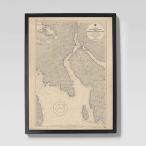 May include: Framed vintage nautical chart in a black frame. The chart shows detailed illustrations of a coastal area, with depth lines and navigational markers. The map is in beige and grey, with text at the top.