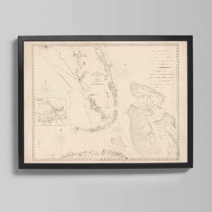 May include: Framed vintage nautical chart in a black frame. The chart displays detailed coastlines, compass roses, and text, in beige and grey tones. The map is a detailed depiction of a coastal area, likely for navigation purposes.