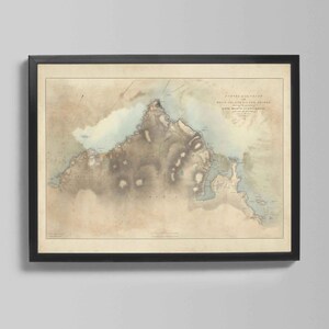 May include: A framed vintage map print in a black frame. The map depicts a detailed illustration of a coastal region, with landmasses in shades of brown and blue. The map has text at the top.