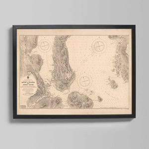 May include: A framed vintage nautical chart, depicting a coastal area with detailed illustrations of land, water depths, and navigational markers. The chart is in sepia tones, with text in the lower center. The frame is black.