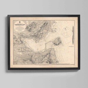 May include: Framed vintage nautical chart of Campbelton Loch, featuring detailed topographical and hydrographical information. The map is in shades of beige and gray, with a black frame. The chart includes text and symbols.