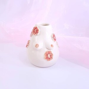 May include: White ceramic vase with a unique shape and floral details. The vase has a rounded body with raised flower accents in shades of pink and orange.