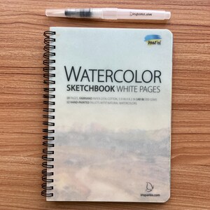 May include: A spiral-bound watercolor sketchbook with white pages, featuring a landscape cover image, the text WATERCOLOR SKETCHBOOK WHITE PAGES, 19 pages, 5.9 in x 8.2 in, 140 lb, and a clear water brush pen with the text insparea.com printed on it.