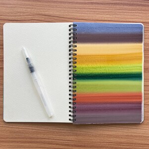 May include: A spiral-bound notebook with a blank cream-colored page on the left and a right-hand page featuring horizontal watercolor stripes in shades of blue, brown, yellow, green, orange, and purple, accompanied by a clear plastic water brush.