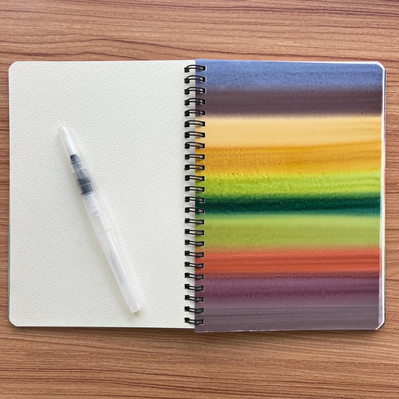 May include: A spiral-bound notebook with a blank cream-colored page on the left and a right-hand page featuring horizontal watercolor stripes in shades of blue, brown, yellow, green, orange, and purple, accompanied by a clear plastic water brush.