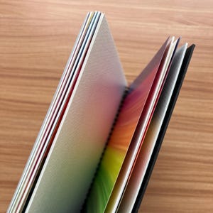 May include: A spiral-bound notebook with a black cover and multiple pages featuring a vibrant rainbow gradient design. The paper edges show a spectrum of colors including red, orange, yellow, green, blue, and violet, with a textured surface.