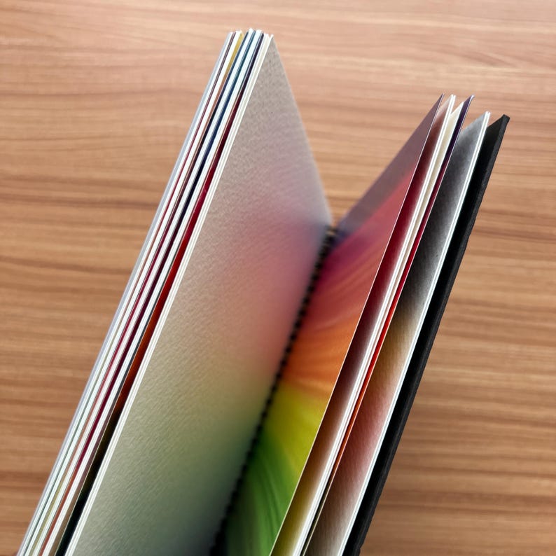 May include: A spiral-bound notebook with a black cover and multiple pages featuring a vibrant rainbow gradient design. The paper edges show a spectrum of colors including red, orange, yellow, green, blue, and violet, with a textured surface.