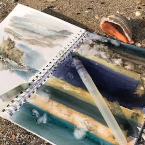 May include: A spiral-bound sketchbook featuring watercolor landscape paintings in shades of blue, green, and tan, with a clear plastic water brush pen resting on the open pages. The artwork depicts abstract coastal scenes with textured paint applications.