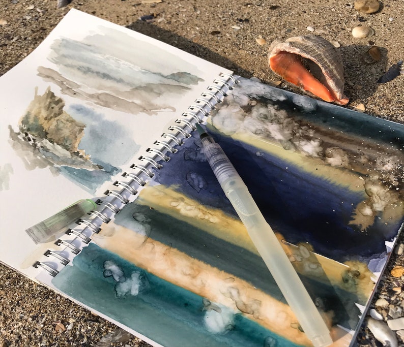 May include: A spiral-bound sketchbook featuring watercolor landscape paintings in shades of blue, green, and tan, with a clear plastic water brush pen resting on the open pages. The artwork depicts abstract coastal scenes with textured paint applications.