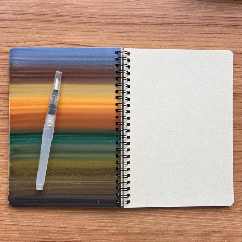 May include: A spiral-bound sketchbook with a cover featuring horizontal bands of blue, brown, orange, teal, and olive green. A clear plastic water brush pen with a white tip rests on the left side of the open notebook, which has blank pages.