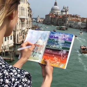 May include: A spiral-bound sketchbook held open, displaying a watercolor painting of a domed building on the left page and a vibrant, multi-colored abstract wash on the right page, being painted with a clear plastic water brush pen.