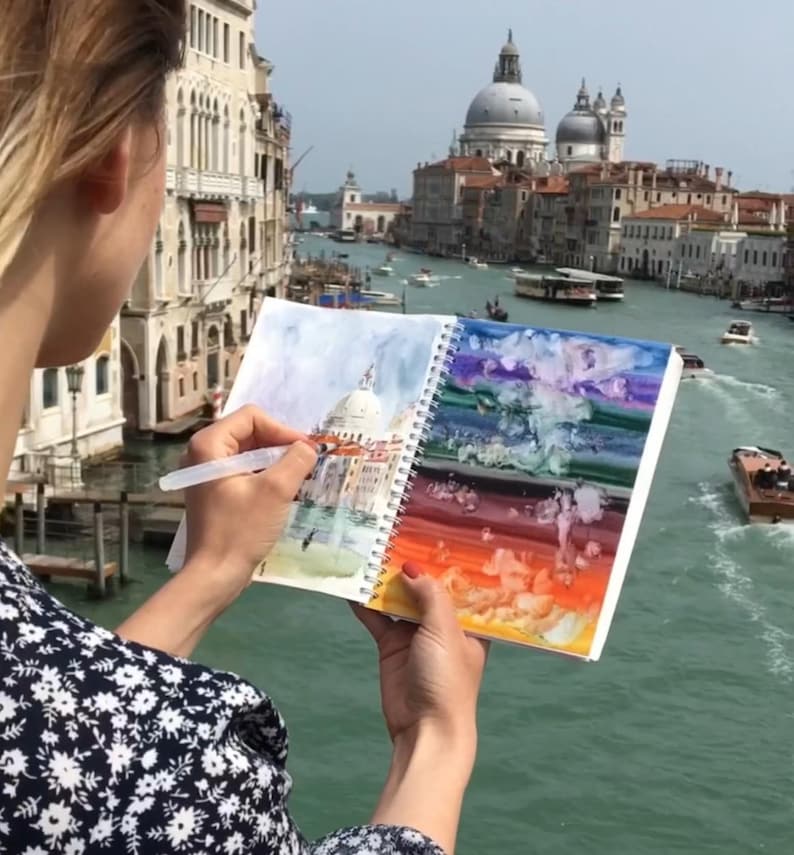 May include: A spiral-bound sketchbook held open, displaying a watercolor painting of a domed building on the left page and a vibrant, multi-colored abstract wash on the right page, being painted with a clear plastic water brush pen.