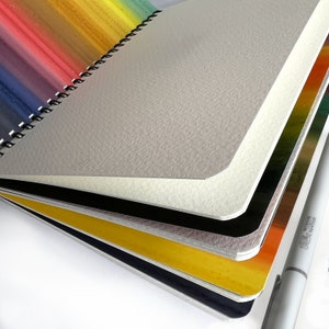 May include: A spiral-bound sketchbook with thick, textured off-white paper pages. The cover features a vibrant rainbow gradient design. A white Winsor & Newton marker is positioned alongside the sketchbook, which has rounded corners on its pages.