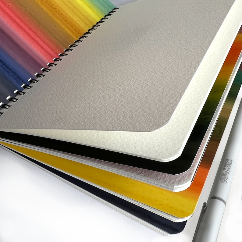 May include: A spiral-bound sketchbook with thick, textured off-white paper pages. The cover features a vibrant rainbow gradient design. A white Winsor & Newton marker is positioned alongside the sketchbook, which has rounded corners on its pages.