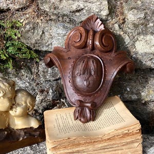 Könnte beinhalten: Eine dunkelbraune, geschnitzte Wandplatte aus Holz mit einem zentralen ovalen Blumenmotiv, platziert hinter einem Stapel gealterter, vergilbter Papierseiten und einer cremefarbenen Skulptur zweier Figuren mit sich berührenden Köpfen.