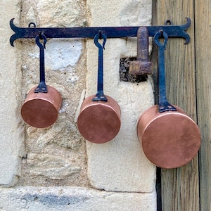 May include: Three small copper pots with black handles hanging from a wrought iron bar. The bar is mounted on a stone wall and a wooden door. The pots are round and have a shiny, reflective surface.