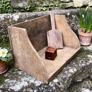 Puede incluir: Un rústico recipiente de madera para pan con una pequeña cuchara de madera y un cubo marrón. El recipiente está sobre una superficie de piedra con un muro de piedra de fondo. Dos plantas en maceta con flores blancas están cerca.