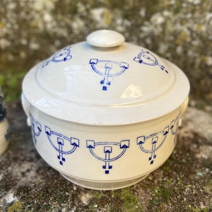 May include: A white ceramic lidded serving dish with blue geometric patterns. The dish has two small handles and a round knob on the lid. A small decorative Christmas tree is to the left of the dish.
