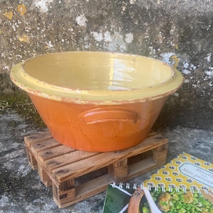 Könnte beinhalten: Eine orangefarbene und gelbe Keramikschüssel mit Griffen, die auf einer kleinen Holzpalette steht. Ein Kochbuch mit grün-weißem Einband und einem Foto von Essen liegt neben der Schüssel. Die Schüssel hat einen breiten Rand und ein glattes Inneres.