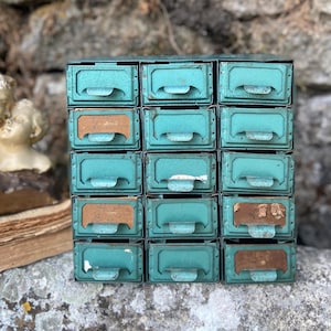 Puede incluir: Un gabinete de almacenamiento de metal color verde azulado con quince cajones individuales dispuestos en una cuadrícula de tres columnas y cinco filas. Cada cajón tiene un asa curva y un portaetiquetas, mostrando signos de desgaste y pintura.
