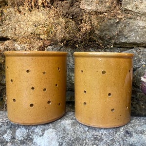 May include: Two cylindrical mustard-yellow ceramic crocks featuring evenly spaced circular ventilation holes throughout the body. The containers have a smooth, glazed finish and a flat base, designed for storage with an open top rim.