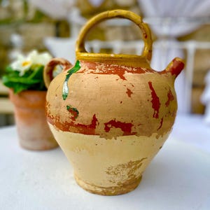 May include: A rustic, hand-painted ceramic pitcher with a handle and spout. The pitcher features a beige base with red and yellow accents, and a green drip. A small terracotta pot with white flowers is in the background.
