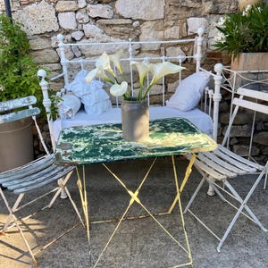 May include: A garden scene with a distressed green and yellow metal table and chairs. A vase of white calla lilies sits on the table. A white wrought iron bench with pillows is in the background.