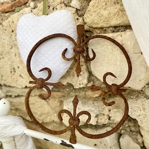 May include: A rusty brown metal decorative wall hanging with a quatrefoil design. The metalwork is mounted on a stone wall, with a white heart-shaped pillow in the background. The pillow is made of quilted fabric.