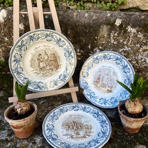 May include: Three antique ceramic plates with blue floral borders and sepia-toned historical scenes. The plates are displayed on a small wooden easel with two potted plants in terracotta pots. The plates are a decorative item.